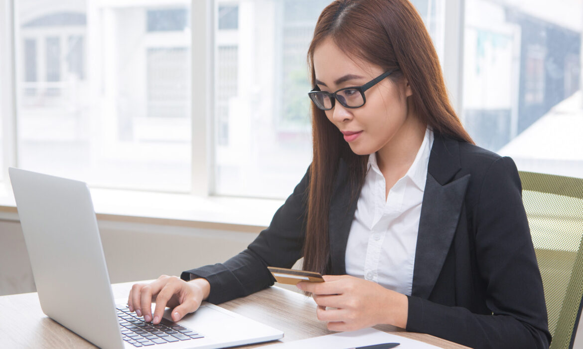 Closeup of content Asian business woman working on laptop computer, holding credit card and doing online banking at office desk with window and houses view outside in background