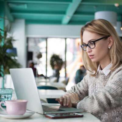 Concentrated female student preparing for exam in cafe. Busy young businesswoman using laptop to work. She reading business article. Lifestyle concept